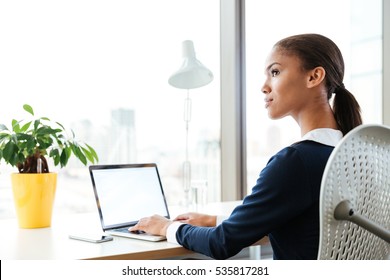 Afro Business Woman In Dress Sitting By The Table Near The Window With Laptop And Looking Aside In Office