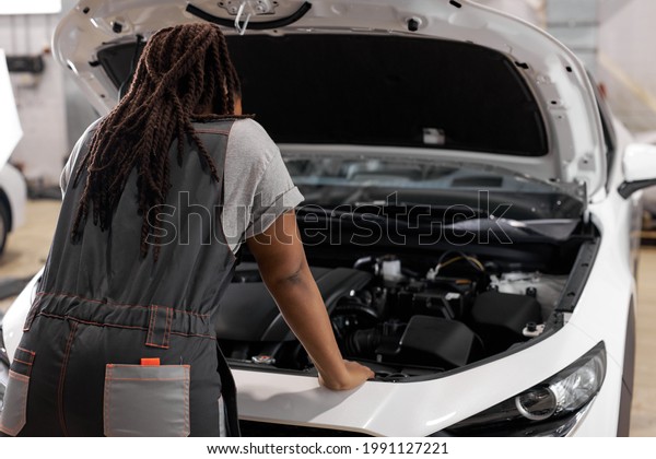 Afro Car mechanic woman is examining under hood of\
car at repair garage, wearing overalls, looking confident and\
concentrated. Rear view on female trying to solve the problem of\
inoperative engine
