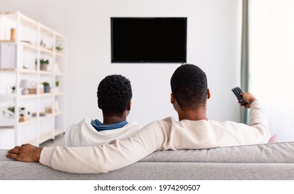 Afro Family Watching TV Sitting On Couch At Home, Rear View. Couple Enjoying Movie Switching Channel Pointing Remote Control At Black Flatscreen Plasma Set On Wall In Living Room. Selective Focus