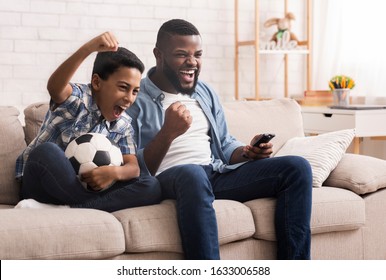 Afro Father And Son Soccer Fans Watching Football On Tv At Home And Emotionaly Cheering, Sitting With Soccer Ball On Couch In Living Room