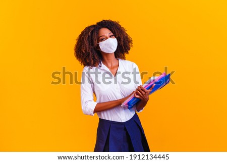 Afro girl holding books on yellow background wearing protective mask. Return to classes by quarantine