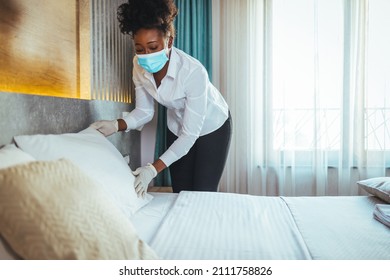 Afro Maid Working At A Hotel Doing Room Disinfection Wearing A Face Mask In Time Of COVID-19 Pandemic. Female Maid With Medical Mask Changing Bed Linen On The Bed