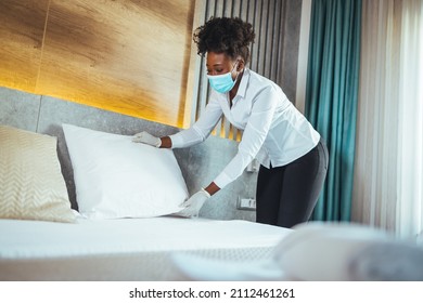 Afro Maid Working At A Hotel Doing Room Disinfection Wearing A Face Mask In Time Of COVID-19 Pandemic. Female Maid With Medical Mask Changing Bed Linen On The Bed