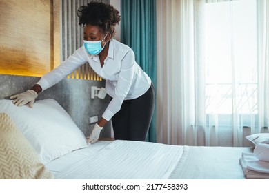 Afro Maid Working At A Hotel Doing Room Disinfection Wearing A Face Mask In Time Of COVID-19 Pandemic. Female Maid With Medical Mask Changing Bed Linen On The Bed