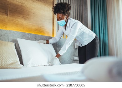 Afro Maid Working At A Hotel Doing Room Disinfection Wearing A Face Mask In Time Of COVID-19 Pandemic. Female Maid With Medical Mask Changing Bed Linen On The Bed