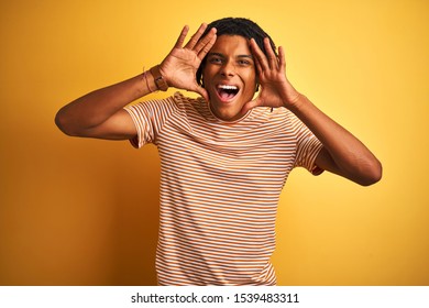 Afro man with dreadlocks wearing striped t-shirt standing over isolated yellow background Smiling cheerful playing peek a boo with hands showing face. Surprised and exited