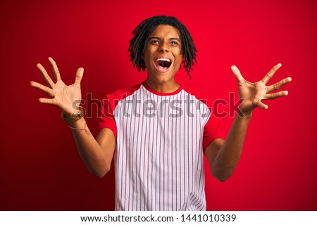 Afro man with dreadlocks wearing striped t-shirt standing over isolated red background celebrating crazy and amazed for success with arms raised and open eyes screaming excited. Winner concept