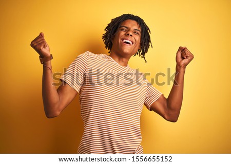Afro man with dreadlocks wearing striped t-shirt standing over isolated yellow background celebrating surprised and amazed for success with arms raised and eyes closed. Winner concept.