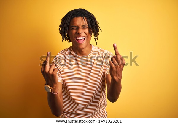 黄色い背景にストライプのtシャツを着たドレッドロックを持つ黒人男性 中指が悪い表情 挑発 失礼な態度を示す わくわくして叫ぶ の写真素材 今すぐ編集