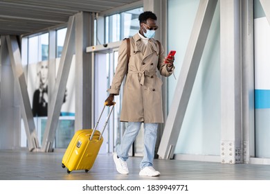 Afro Millennial Man Walking With Yellow Suitcase In Airport Terminal, Using Mobile Phone, Wear Face Mask. Traveler African American Male With Luggage In Railway Station. New Normal, Covid-19 Concept.