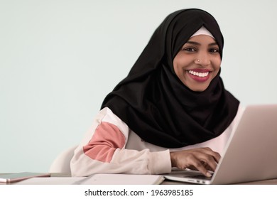 afro muslim woman wearing a hijab sits smiling in her home office and uses a laptop