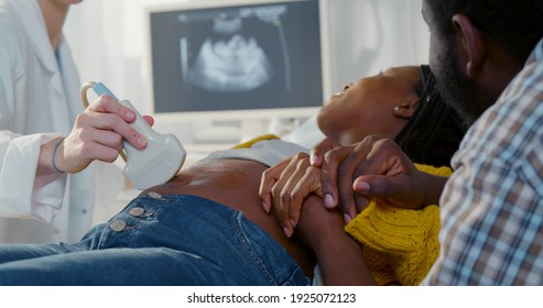Afro pregnant couple smiling looking at sonogram results on monitor while on checkup at gynecologist. African husband holding hand of pregnant wife during ultrasound