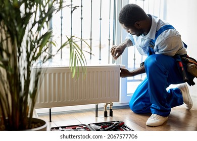 afro repairman in blue overalls using tools while installing or repairing heating radiator. plumber installing heating system in apartment. radiator installation, plumbing works and home renovation