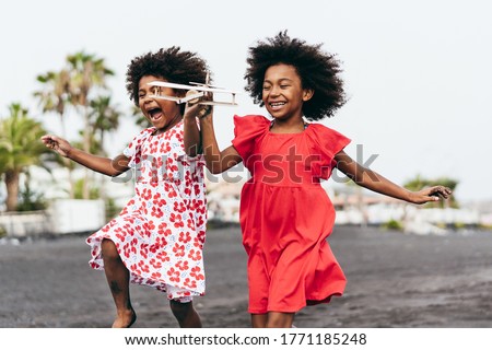 Afro twins sisters running on the beach while playing with wood toy airplane - Youth lifestyle and travel concept - Main focus on right kid face