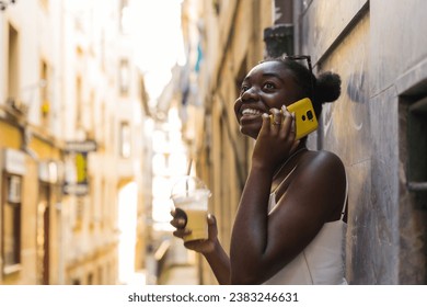 Afro woman leaning on a city wall drinking fresh juice and talking to the mobile - Powered by Shutterstock