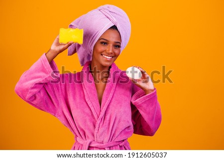 Afro woman with towel on her head and bathrobe holding a sponge and soap on yellow background. Bath concept
