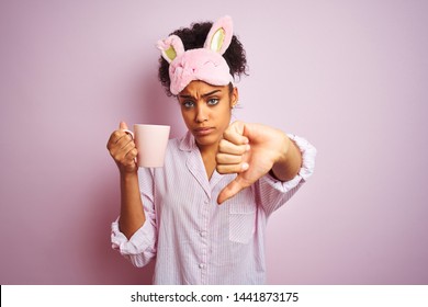 Afro woman wearing pajama and mask drinking a cup of coffee over isolated pink background with angry face, negative sign showing dislike with thumbs down, rejection concept