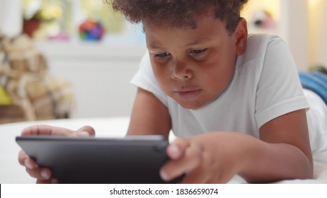 Afro-american boy kid lying on bed watching cartoons on digital tablet. Close up portrait of adorable african child playing game on tablet pc relaxing in bedroom at home
