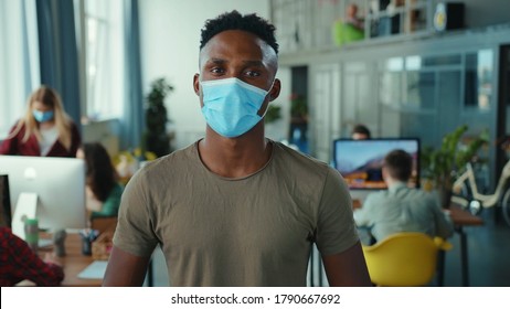 Afro-american Business Young Employee In Respiratory Mask At Open Space Office. Young People On Background Coworking And Discussing Plans. Workspace. Healthcare. Pandemic.