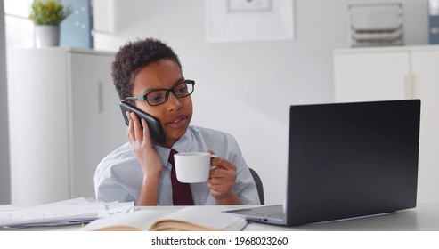 Afro-american Child Boss In Glasses Talking On Smartphone And Drinking Coffee. Portrait Of African Kid Businessman Having Phone Call Sitting At Desk In Office