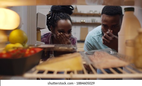 Afro-american Couple Opening Fridge And Wrinkling Face Having Food Spoiled. View From Inside Of African Man And Woman Opening Refrigerator With Stinking Meal