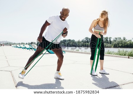 Afroamerican fitness instructor in white t-shirt and black shorts with tape expander doing exercises with women in grey top and black pants on fresh air and sunny weather