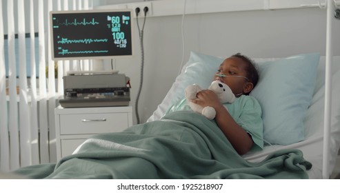 Afro-american Kid Lying In Hospital Bed With Oxygen Tube And Plush Toy. Portrait Of Sick And Sad African Child Patient Resting In Bed At Hospital Ward With Modern Equipment