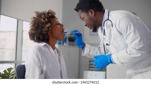 Afro-american Male Doctor Examining Young Woman Throat In Office. Black Man Therapist Using Wooden Stick Checking Sore Throat Of Young Female Patient