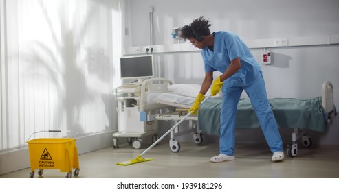 Afro-american Male Janitor In Headphones Washing Floor In Hospital Ward. Young African Man Nurse Listening To Music In Earphones And Cleaning Floor With Mop In Clinic Room