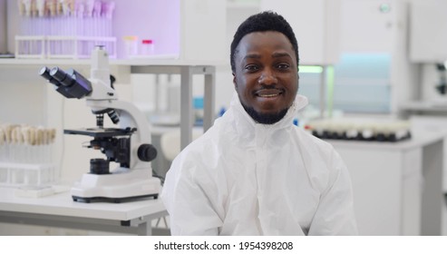 Afro-american Male Scientist In Overall Smiling At Camera In Lab. Portrait Of Young Black Virologist In Safety Uniform Working In Modern Hospital Laboratory