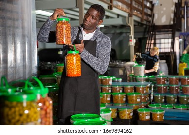 Afro-american male worker stocks plastic containers and cans with olives in warehouse