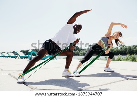 Afroamerican man in white t-shirt and black shorts with tape expander doint exercises with women in grey top and black pants on fresh air and sunny weather