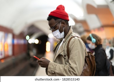 Afro-American millennial man in trench coat, red hat wearing face mask as protection against  covid-19, flu virus, waiting for the train at subway station, using mobile phone. New normal, pandemic. - Powered by Shutterstock