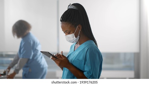 Afro-american Nurse Wearing Safety Mask Using Digital Tablet In Hospital Corridor. African Female Surgeon In Scrubs And Protective Mask Reading Report On Tablet Pc In Clinic Hallway