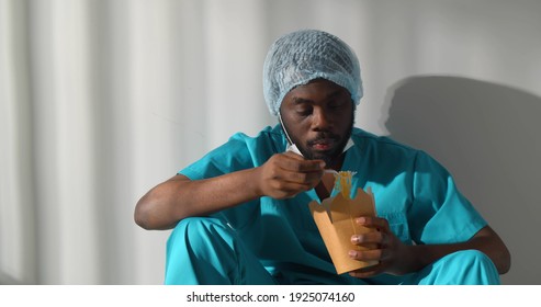 Afro-american Surgeon In Scrubs And Safety Mask Eating Takeaway Lunch On Floor In Hallway. African Man Nurse Having Break And Eating Meal Sitting On Floor In Corridor