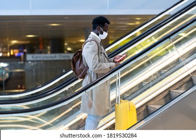 Afro-American traveler man with yellow suitcase stands on escalator in airport terminal, wear face medical mask to protect yourself from contact with flu virus, pandemic covid-19. New normal concept  - Powered by Shutterstock