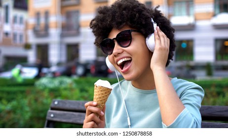 Afro-american Woman Enjoying Music In Headphones, Eating Ice-cream On Bench