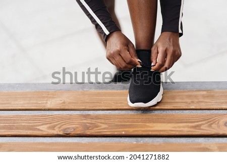 Afro-American woman hand tying sport black shoe lace while holding foot on a wood bench outdoors. Multiethnic women having a fitness workout break.