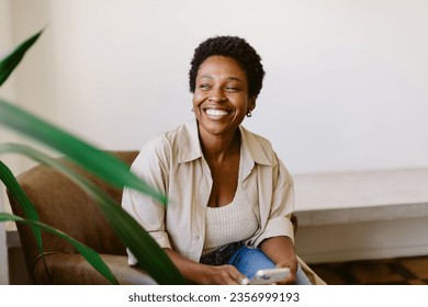 Afro-haired woman sits on a couch in her home, smiling and holding a smartphone. She looks away, enjoying a moment of candid relaxation and social media browsing. - Powered by Shutterstock