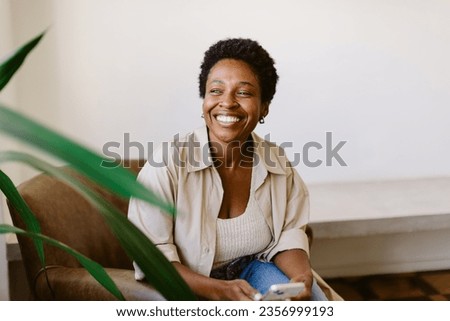Afro-haired woman sits on a couch in her home, smiling and holding a smartphone. She looks away, enjoying a moment of candid relaxation and social media browsing.