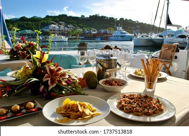 Aft deck dining table set for a cocktail party with appetizers, wine glasses and wine bucket in late afternoon at Falmouth Harbour with Antigua Yacht Club in background.