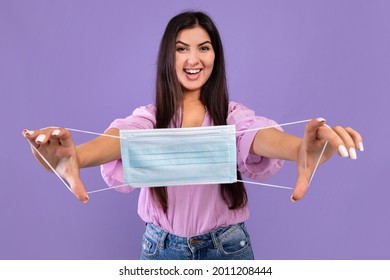 After Covid. Excited Armenian Woman Holding Medical Face Mask In Outstretched Hands And Smiling At Camera On Purple Studio Background. Happy Lady Free From Coronavirus Protection