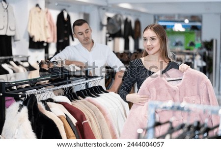After inspection of stores assortment, girl chooses fur coat on hangers and closely examines its details, feels texture of fabric. Buyer thoughtfully goes through folds touch of overcoat fabric