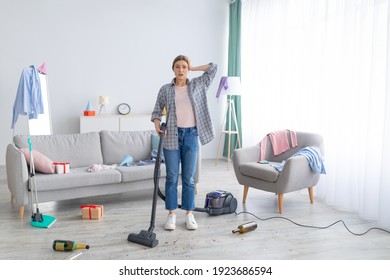 After Party Chaos. Dismayed Millennial Woman Standing In Her Messy Apartment With Vacuum Cleaner, Holding Head In Terror, Copy Space. Terrified Female Student Cleaning Dirty University Dorm Room