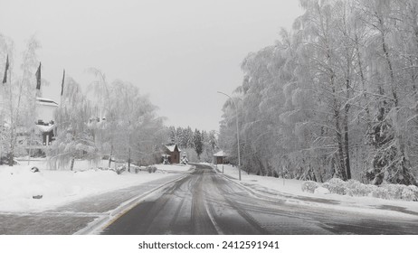 After a snowfall, snow lies on the road, tree branches and roofs of houses. Along the street there are houses, trees, and poles with streetlights. The roadway and sidewalk are cleaned. Overcast - Powered by Shutterstock