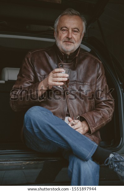 After the trip. An\
elderly man with a gray beard in a leather jacket holds in his hand\
a glass of whiskey against the background of an open trunk of a car\
in a garage\
