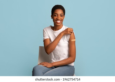 After Vaccination Concept. Portrait Of Vaccinated Black Woman Showing Arm After Coronavirus Vaccine Injection, Positive Lady Sitting With Rolled Up Sleeve Over Blue Background, Smiling To Camera