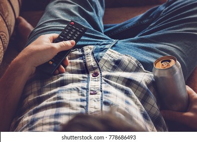 After work a guy lying on sofa, drinking a cold beer and watching sport tv channel. Hand holding a aluminum can and remote control. Man's resting time at home concept. 
