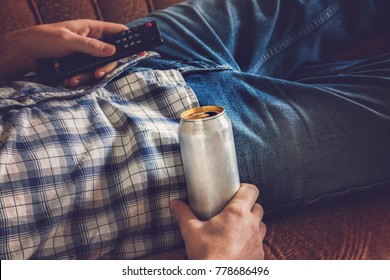 After Work A Guy Lying On Sofa, Drinking A Cold Beer And Watching Sport Tv Channel. Hand Holding A Aluminum Can And Remote Control. Man's Resting Time At Home Concept. 
