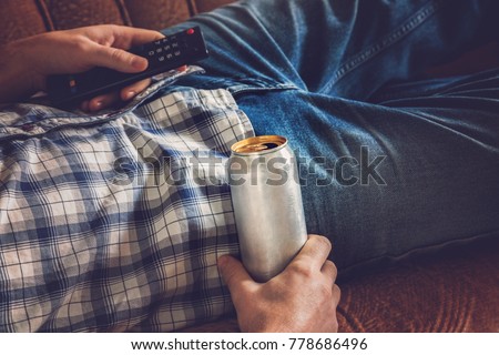 After work a guy lying on sofa, drinking a cold beer and watching sport tv channel. Hand holding a aluminum can and remote control. Man's resting time at home concept. 
